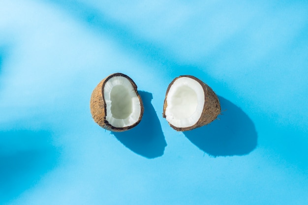 Unterbrochene Kokosnuss auf einer blauen Oberfläche unter natürlichem Licht mit Schatten. Hartes Licht. Konzept der Ernährung, gesunde Ernährung, Erholung in den Tropen, Urlaub und Reisen, Vitamine. Flachgelegt, Draufsicht.