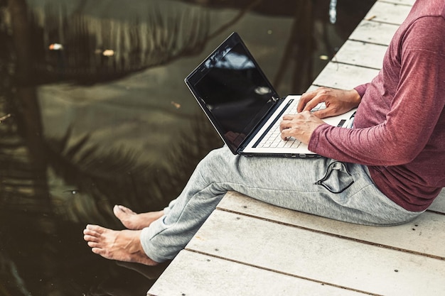 Foto unterer teil eines mannes, der einen laptop benutzt, während er auf dem pier sitzt