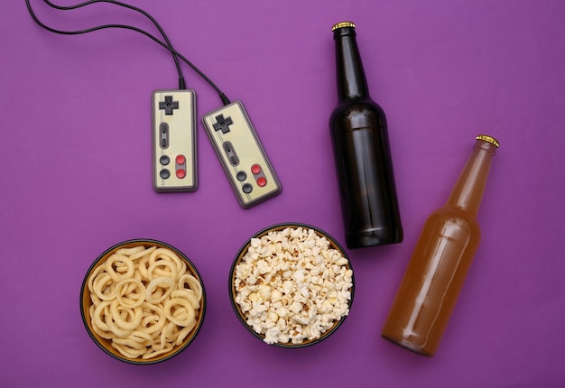 Unterhaltungszeit. Gamepads und Snackschüssel, Bierflaschen auf violettem Hintergrund. Ansicht von oben