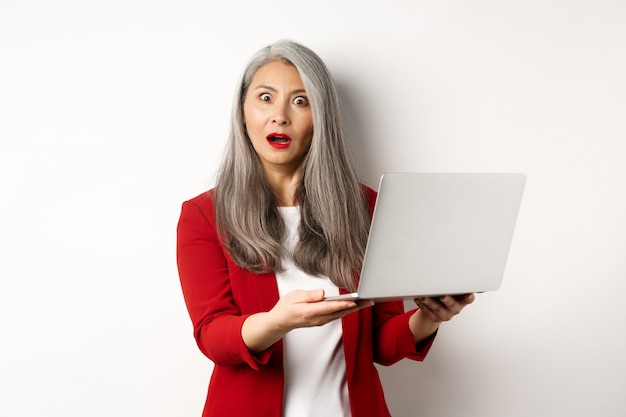 Unternehmen. Beeindruckte asiatische alte Frau, die am Laptop arbeitet, überrascht in die Kamera starrt, stehend auf weißem Hintergrund.