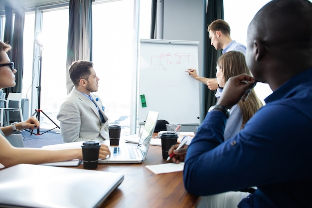 Foto unternehmensfortschritt diskutieren. zuversichtlich junger mann, der nahe whiteboard steht und grafik zeigt, während seine kollegen am schreibtisch sitzen.