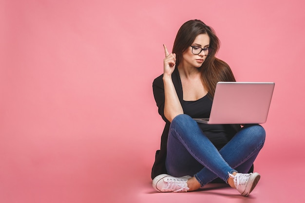 Unternehmenskonzept. Porträt der glücklichen Frau im lässigen Sitzen auf Boden in der Lotushaltung und im Halten des Laptops lokalisiert über rosa Hintergrund