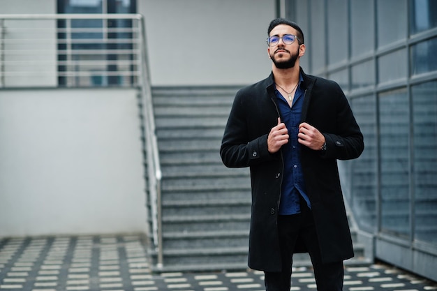 Unternehmer aus dem Nahen Osten tragen einen schwarzen Mantel und eine blaue Hemdbrille gegen ein Bürogebäude, das auf Treppen geht