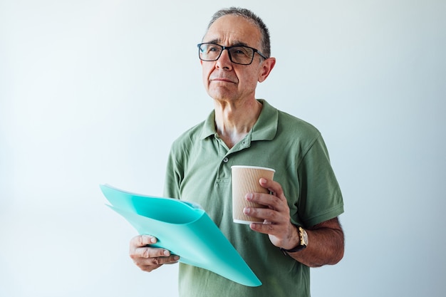 Unternehmer pensionierter Mann, der ein grünes Hemd und eine Brille trägt, einen Ordner mit einem Bericht und einem Kaffee hält, denkt