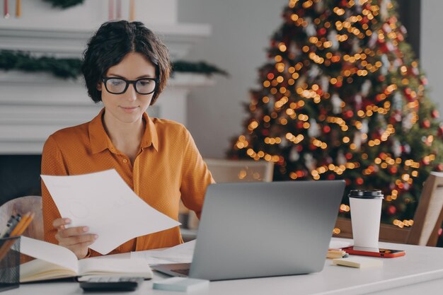 Unternehmerin arbeitet zu Weihnachten online am Laptop im Büro mit Weihnachtsbaum im Hintergrund