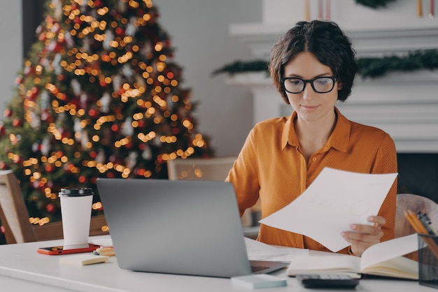 Unternehmerin arbeitet zu Weihnachten online am Laptop im Büro mit Weihnachtsbaum im Hintergrund