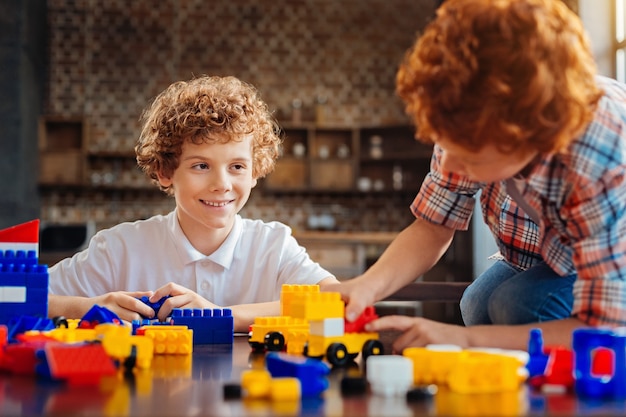 Unterstützende hochstimmung. selektiver fokus auf einen lockigen jungen, der breit lächelt, während er an einem tisch sitzt und seinen kleinen bruder ansieht, während beide mit einem baukasten spielen.