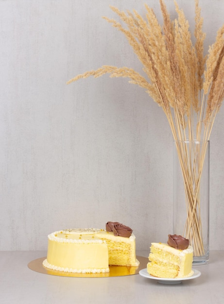 Untertasse ist ein geschnittenes Stück Kuchen aus einem gelben Sahnekuchen mit Schokoladenrosen und einem Strauß getrockneter Schilfblumen in einer Glasvase auf grauem Hintergrund mit Kopierraum
