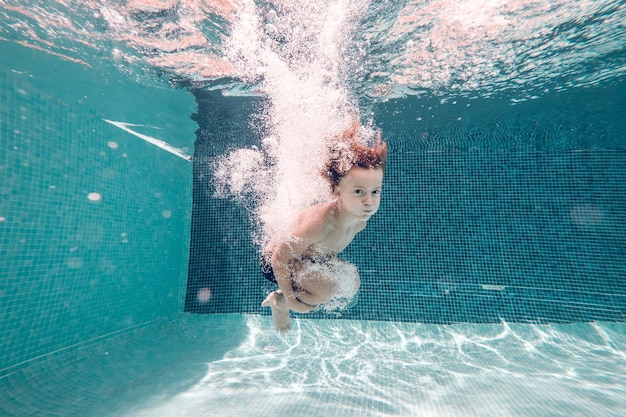 Unterwasseransicht von barfußem Kind mit nacktem Rumpf, das Schwimmhose trägt, springt und taucht in klarem, durchsichtigem Wasser, umarmt Knie und schaut in die Kamera