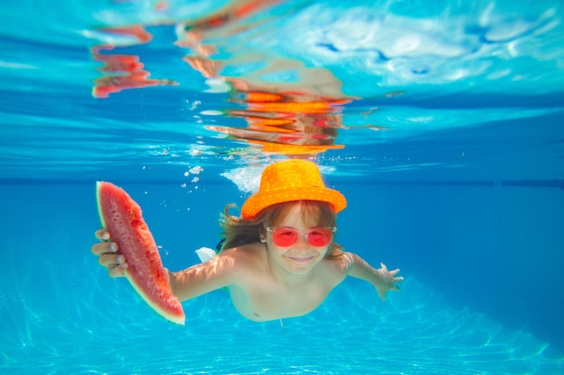 Unterwasserjunge hält Wassermelone im Schwimmbad Süßer Junge, der im Pool unter Wasser schwimmt