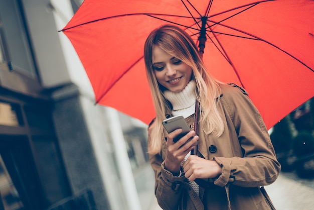 Unterwegs eine Nachricht eingeben. Attraktive junge lächelnde Frau, die Regenschirm trägt und ihr Handy ansieht, während sie auf der Straße steht