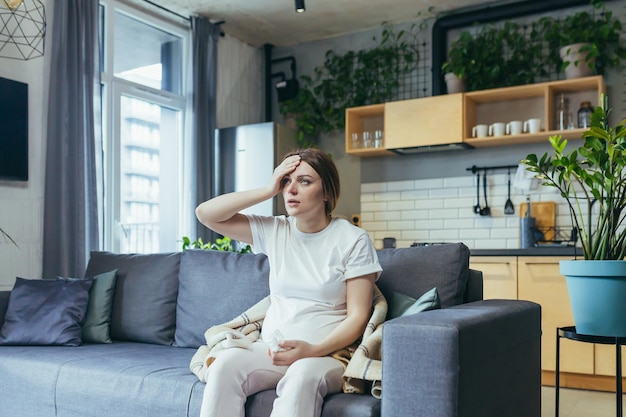 Unwohlsein Eine schwangere Frau ist krank, hat Kopfschmerzen und hält ihre Hand hinter dem Kopf Sitzt zu Hause in Heimkleidung auf der Couch