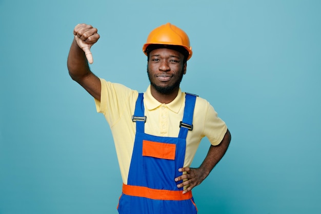 unzufrieden zeigt Daumen nach unten und legt die Hand auf die Hüften junger afroamerikanischer Baumeister in Uniform isoliert auf blauem Hintergrund