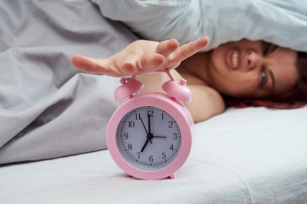 Unzufriedene Frau, die im Bett liegt und den Wecker am frühen Morgen stoppt.