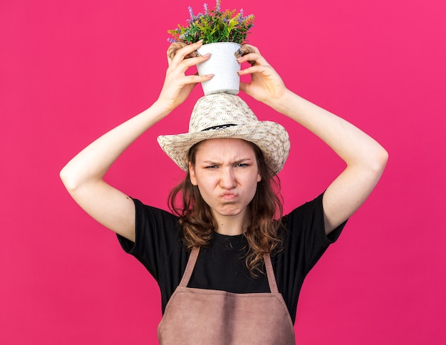 Unzufriedene junge Gärtnerin mit Gartenhut, die Blume im Blumentopf auf dem Kopf hält