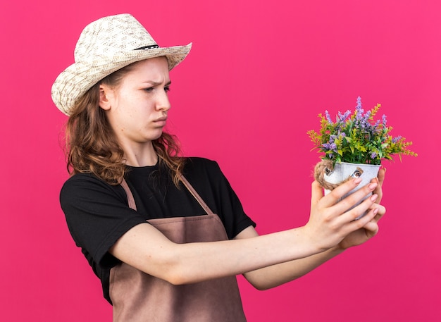 Unzufriedene junge Gärtnerin mit Gartenhut, die Blume im Blumentopf hält und betrachtet