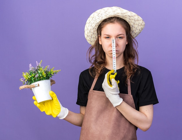 Unzufriedene junge Gärtnerin mit Gartenhut mit Handschuhen, die Blume im Blumentopf mit Maßband hält