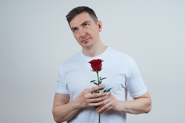 Unzufriedener erwachsener Mann in einem weißen T-Shirt mit einer roten Rose in seinen Händen auf einem grauen Hintergrund. Fröhlichen Valentinstag.