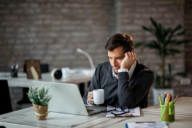 Unzufriedener Geschäftsmann trinkt Kaffee und liest eine E-Mail auf dem Computer, während er im Büro arbeitet