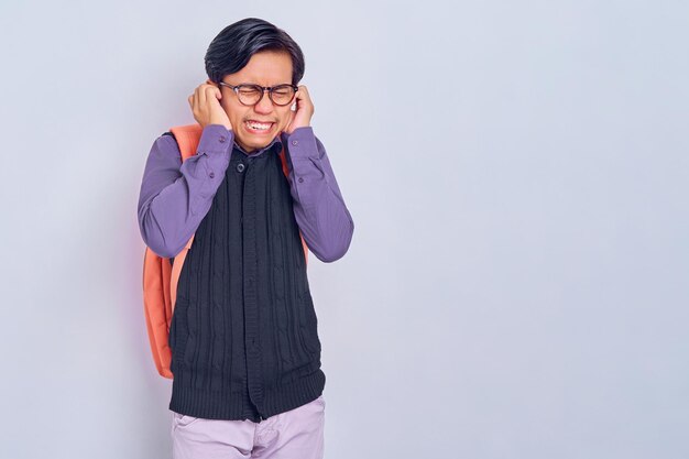 Unzufriedener junger asiatischer Student in Freizeitkleidung mit Rucksack, der die Ohren mit der Hand bedeckt, die auf grauem Hintergrund isoliert ist Konzept der High School University College