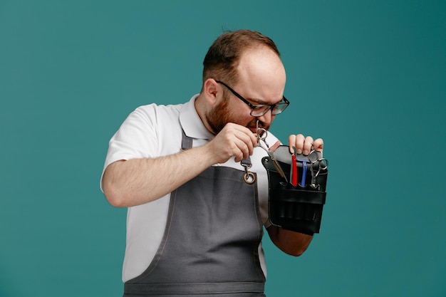 Unzufriedener junger Friseur in Uniform und Brille, der die Friseurtasche hält und betrachtet, die eine Schere hineinsteckt, isoliert auf blauem Hintergrund