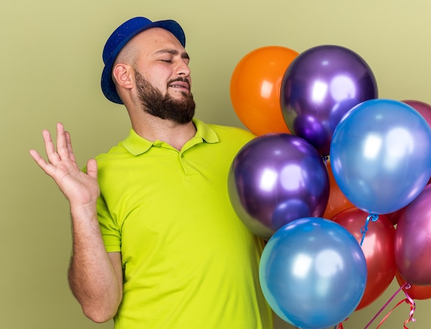 Foto unzufriedener junger mann mit partyhut, der luftballons hält und betrachtet, die hand isoliert auf olivgrüner wand ausbreiten?