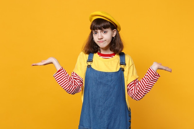 Unzufriedener Mädchen-Teenager in französischer Baskenmütze, sich ausbreitendes Denim-Sommerkleid, Hände zur Seite zeigend, isoliert auf gelbem Wandhintergrund im Studio. Menschen aufrichtige Emotionen, Lifestyle-Konzept. Mock-up-Kopienbereich.