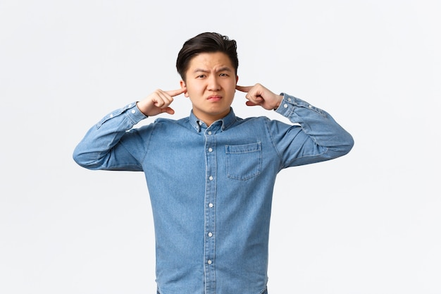 Unzufriedener und belästigter asiatischer männlicher Student, der sich über lauten Nachbarn beschwert, eine Grimasse verzieht, die Ohren mit den Fingern schließt und genervt aussieht, durch lautes Geräusch gestört wird, weißer Hintergrund steht.