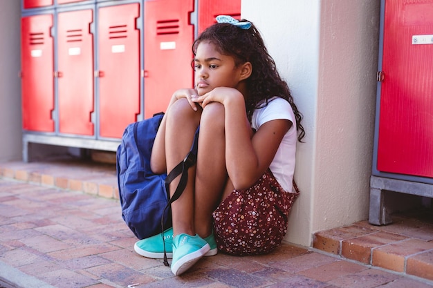 Unzufriedenes afrikanisch-amerikanisches Schulmädchen sitzt mit Schultasche neben Schließfächern im Schulkorridor