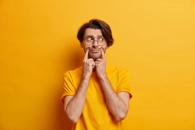 Unzufriedenheit unrasierter europäischer Mann zwingt Lächeln hält Zeigefinger in der Nähe von Lippenwinkeln trägt runde Brillen lässiges T-Shirt isoliert über gelber Wand. Hipster-Typ gibt vor, glücklich zu sein