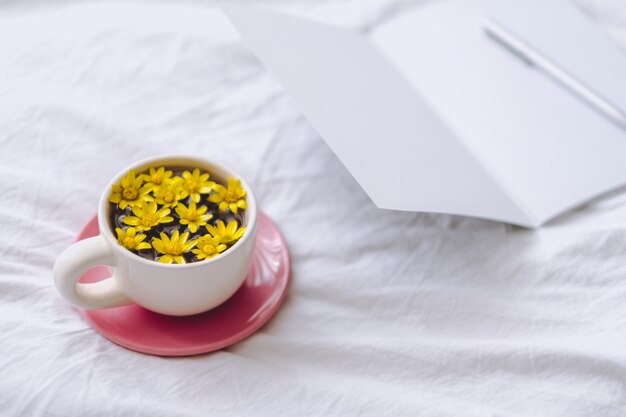 Ð¡up con flores amarillas en el interior, sobre una cama blanca con bloc de notas y bolígrafo.