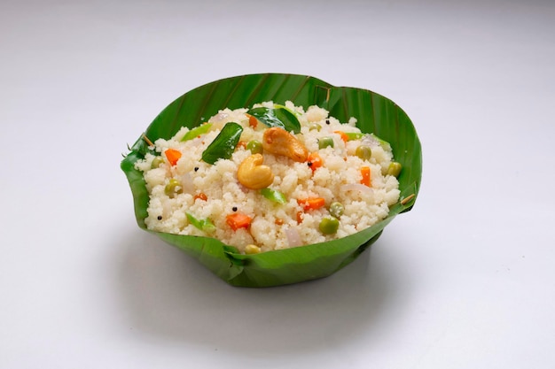 Upma feito de samolina ou rava, item de café da manhã do sul da Índia que é lindamente organizado em uma tigela feita de folha de bananeira e guarnecido com castanha de caju frita em guardanapo com fundo texturizado branco.