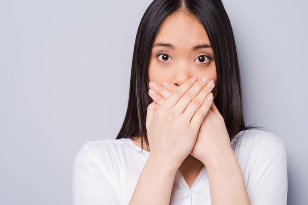 ¡UPS! Mujer asiática joven sorprendida que cubre la boca con las manos y mirando a la cámara mientras está de pie contra el fondo gris