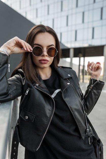 Urban schönes junges cooles Mädchen mit dem sexy Schönheitsgesicht in modischer runder Sonnenbrille mit stylischer schwarzer Lederjacke und Kleid mit Handtasche geht auf die Straße