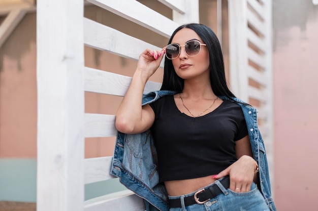 Urban stilvolle junge Frau Hipster in einem trendigen schwarzen Top in einer modischen Jeansjacke in einem Rock in dunkler Sonnenbrille posiert in der Nähe einer weißen Holzwand in der Stadt. Attraktives Mädchenmodel im Freien.