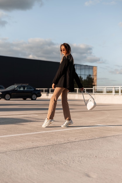 Urban trendige junge Frau mit Tasche in stilvoller Freizeitkleidung und Schuhen geht zum Einkaufszentrum. Modernes Mädchen posiert auf dem Parkplatz im Freien an einem sonnigen Sommertag. Elegantes Aussehen.