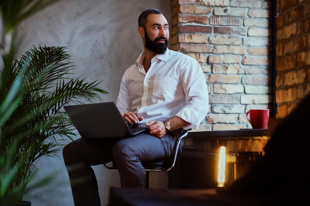 Urbaner bärtiger Mann trinkt Kaffee und benutzt einen Laptop in einem Raum mit Loft-Interieur.