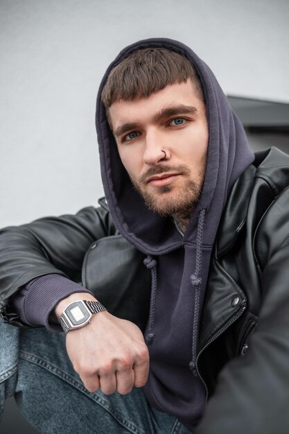 Urbanes männliches Porträt eines gutaussehenden, stilvollen Hipsters mit stylischem Freizeitoutfit mit Lederjacke, Hoodie und Jeans mit Uhr auf der Straße