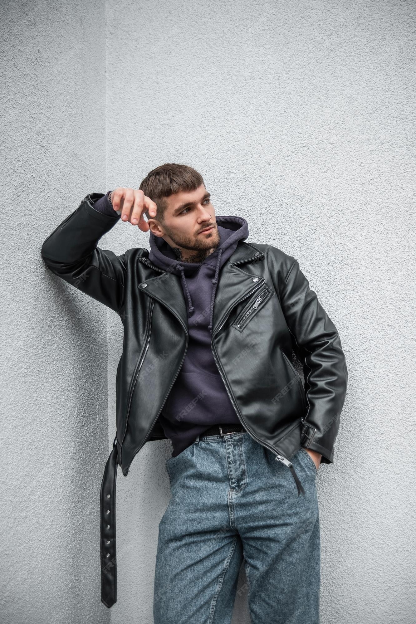 Urbano de moda, apuesto hombre hipster con chaqueta de cuero rock de moda, con capucha y jeans, se encuentra cerca de una pared blanca en la calle | Foto Premium