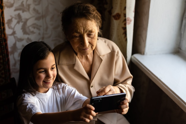 Urgroßmutter sitzt bei Urenkelin und schaut ins Smartphone. Großmutter und Kind machen ein Selfie auf einem Smartphone. Großmutter mit ihrer Enkelin schauen ins Telefon.