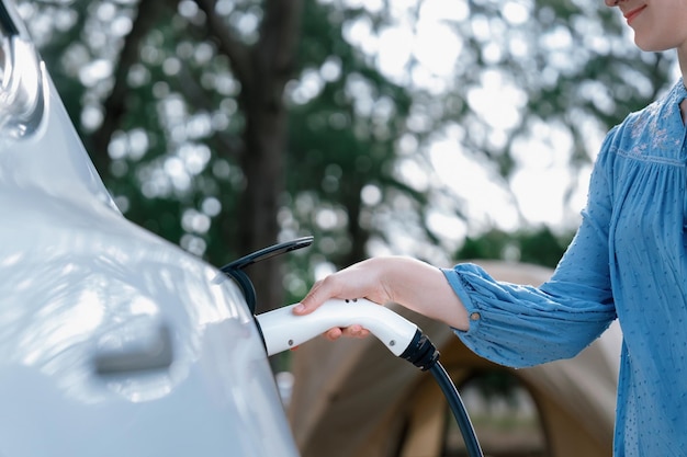 Urlaub auf dem Campingplatz, Reisen in die Natur mit einem Elektroauto. Frau lädt ihr Elektroauto an der Ladestation auf dem Campingplatz auf. Natürliches Reisen und Öko-Auto für eine nachhaltige Umwelt. Perpetual