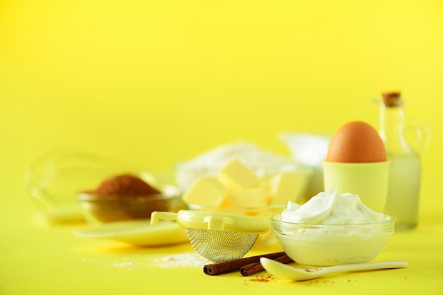 Urlaub Bäckerei Hintergrund. Lebensmittelrahmen - Butter, Zucker, Mehl, Milch, Eier, Öl, Löffel, Nudelholz, Bürste, Schneebesen, Zimt über gelbem Hintergrund.