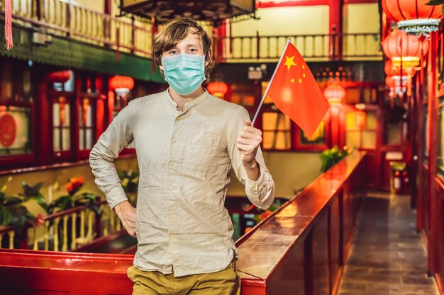 Urlaub in China genießen junger Mann mit chinesischer Flagge in medizinischer Maske auf chinesischem Hintergrund