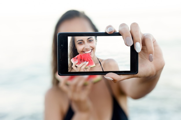 Urlaub, Resort, Tourismuskonzept - junges Mädchen isst frische Wassermelone am Sandstrand. Porträt-Teenager-Mädchen mit einem Smartphone-Gerät, das Selfies von sich selbst im Urlaub gegen blauen Himmel macht