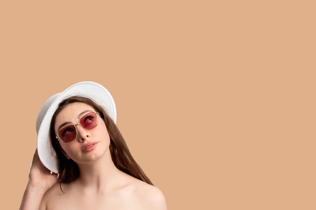 Urlaubsreise Sommeroutfit Stilvolle Frau mit nackten Schultern in weißem Zigeunerhut und Sonnenbrille, die isoliert auf beigem Kopierraum nach oben schaut Sonnenschutzzubehör Reisetour