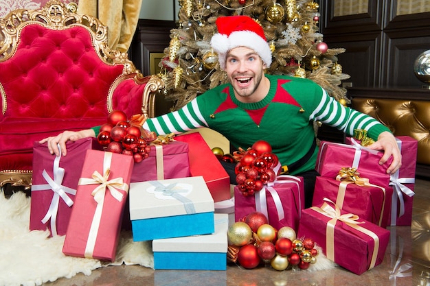 Urlaubsvorbereitung, Feier, Party. Mann im Sankt-Hutlächeln mit Geschenken am Weihnachtsbaum. Frohes neues Jahr, Weihnachten, Vorabend. Macho im Elfenkostüm mit Schachteln. Schenken, Boxtag-Konzept.