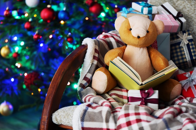 Urso de pelúcia com livro e caixas de presente em uma cadeira de balanço no fundo da árvore de Natal