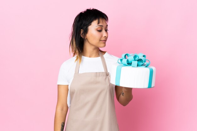 Uruguayischer Konditor, der einen großen Kuchen über lokalisiertem rosa Hintergrund hält, der zur Seite schaut