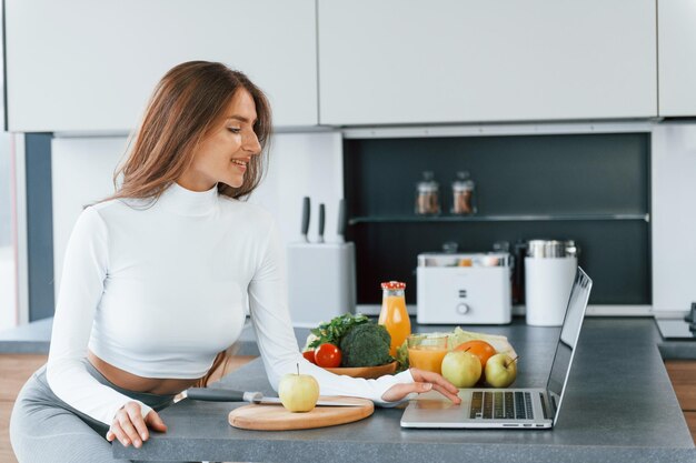 Usa laptop Jovem europeia está dentro de casa na cozinha dentro de casa com comida saudável