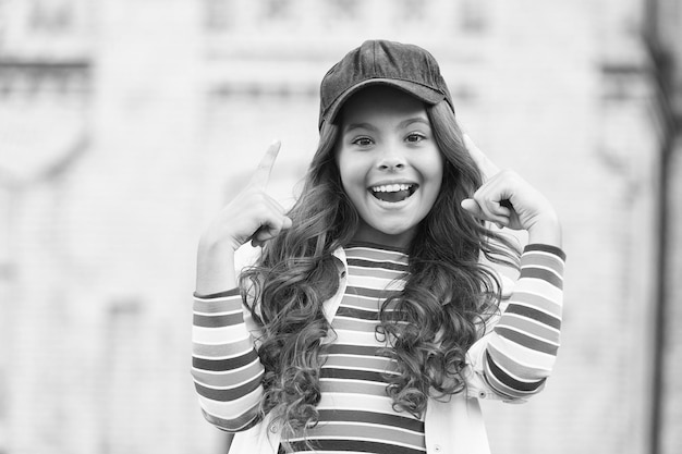 Usa tu cerebro Un niño feliz señala con el dedo al aire libre Un niño pequeño usa una gorra con el pelo largo y ondulado Sonrisa de un niño pequeño con ropa informal Accesorio de moda Estilo moderno Cuidado infantil e infancia